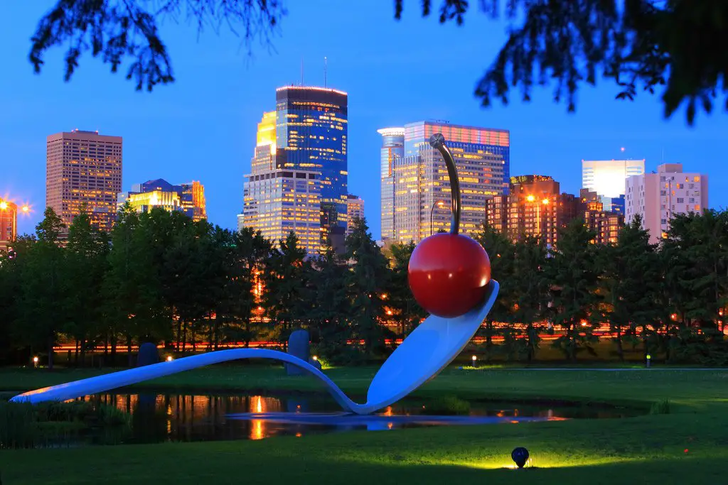 The Minneapolis skyline shines behind the Spoonbridge and Cherry at the Minneapolis Sculpture Garden, part of the Walker Art Center collection. (Photo courtesy of Meet Minneapolis | Spoonbridge and Cherry, Claes Oldenburg and Coosje van Bruggen, 1985-1988, aluminum, stainless steel, paint, 354 x 618 x 162"overall, Collection Walker Art Center, Minneapolis, Gift of Frederick R. Weisman in honor of his parents, William and Mary Weisman, 1988, Art © Claes Oldenburg and Coosje van Bruggen)
