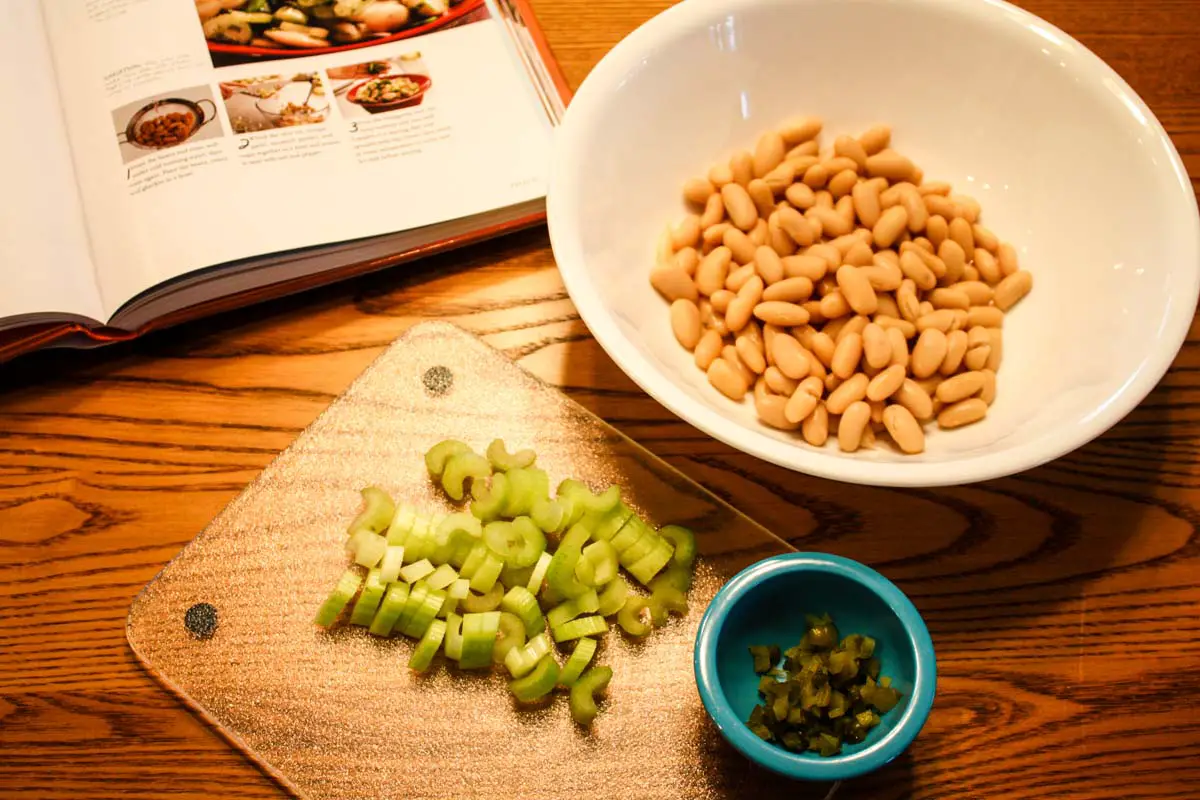 Atop a wooden table sits an open cookbook, white bowl filled with cannellini beans, chopped celery stalks on a clear cutting board, and small blue bowl of finely chopped gherkin pickle