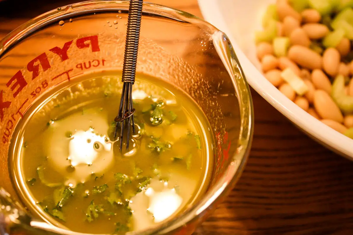 Pyrex glass liquid measuring cup filled with olive oil-based vinaigrette with whisk in mixture and cropped bowl of cannellini beans to right side