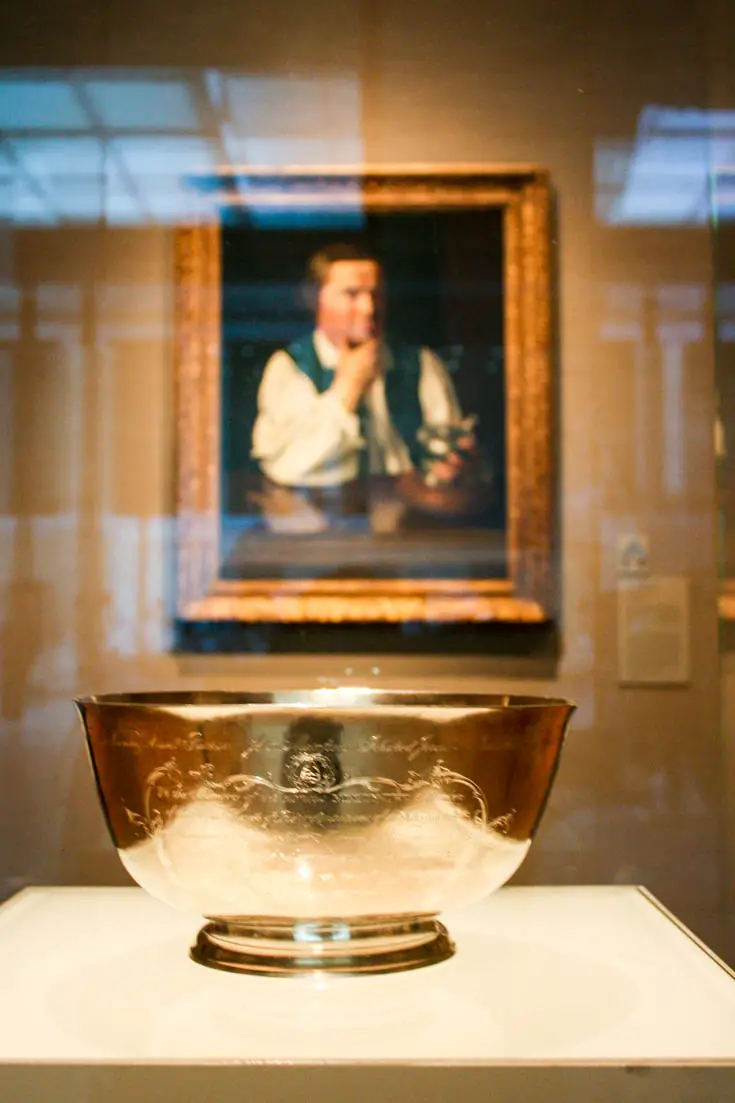 "Sons of Liberty Bowl" by American silversmith Paul Revere with Revere's portrait by John Singleton Copley behind it displayed in the Art of the Americas collection at the Museum of Fine Arts, Boston