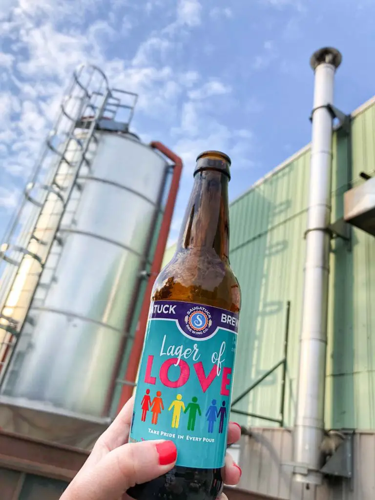 Woman's hand with brightly colored nail polish holds a bottle of Saugatuck Brewing Co. Lager of Love in front of brewery facility and blue sky dotted with fluffy white clouds