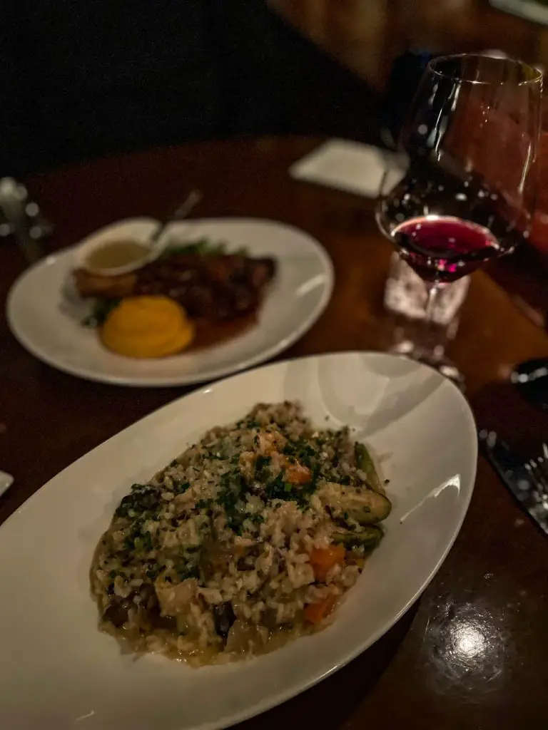 Wild mushroom risotto with lamb shank main dish in the background and a glass of red wine in a dimly lit restaurant setting at Divani in Grand Rapids, Michigan, USA
