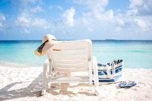 Lounge chair with a floppy beach hat, a beach bag, and flipflops on a white sand beach with aqua water and fluffy white clouds in the sky