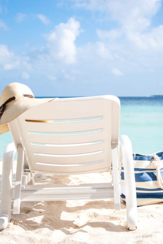 Lounge chair with a floppy beach hat, a beach bag, and flipflops on a white sand beach with aqua water and fluffy white clouds in the sky