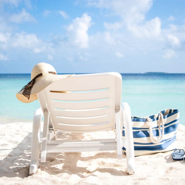 Lounge chair with a floppy beach hat, a beach bag, and flipflops on a white sand beach with aqua water and fluffy white clouds in the sky