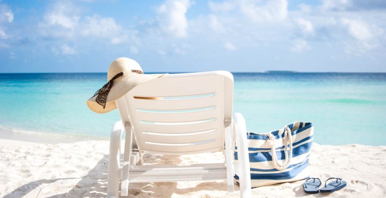 Lounge chair with a floppy beach hat, a beach bag, and flipflops on a white sand beach with aqua water and fluffy white clouds in the sky