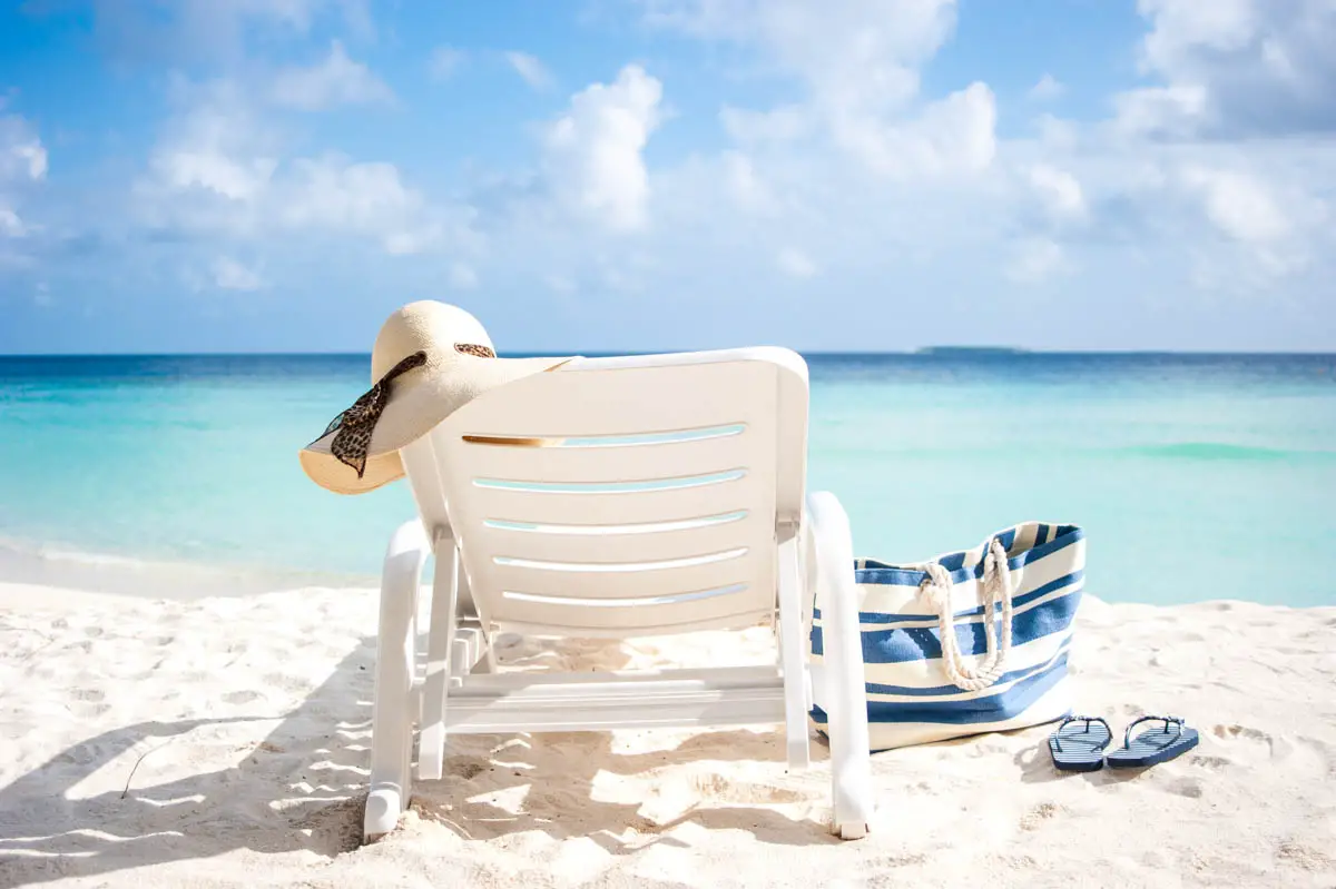 Lounge chair with a floppy beach hat, a beach bag, and flipflops on a white sand beach with aqua water and fluffy white clouds in the sky