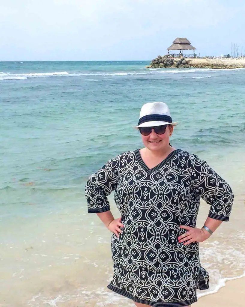 Woman wearing a swimsuit coverup dress, a fedora-style beach hat, and sunglasses stands on the beach in Mexico's Riviera Maya