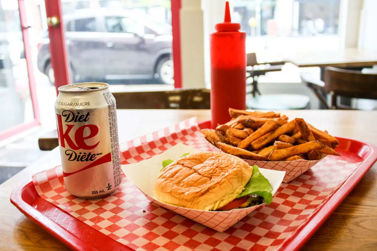 Cheeseburger, fries, ketchup bottle, and Diet Coke can set on a red-and-white checkered paper-lined red tray inside a restaurant with a street view in Toronto's Little Italy