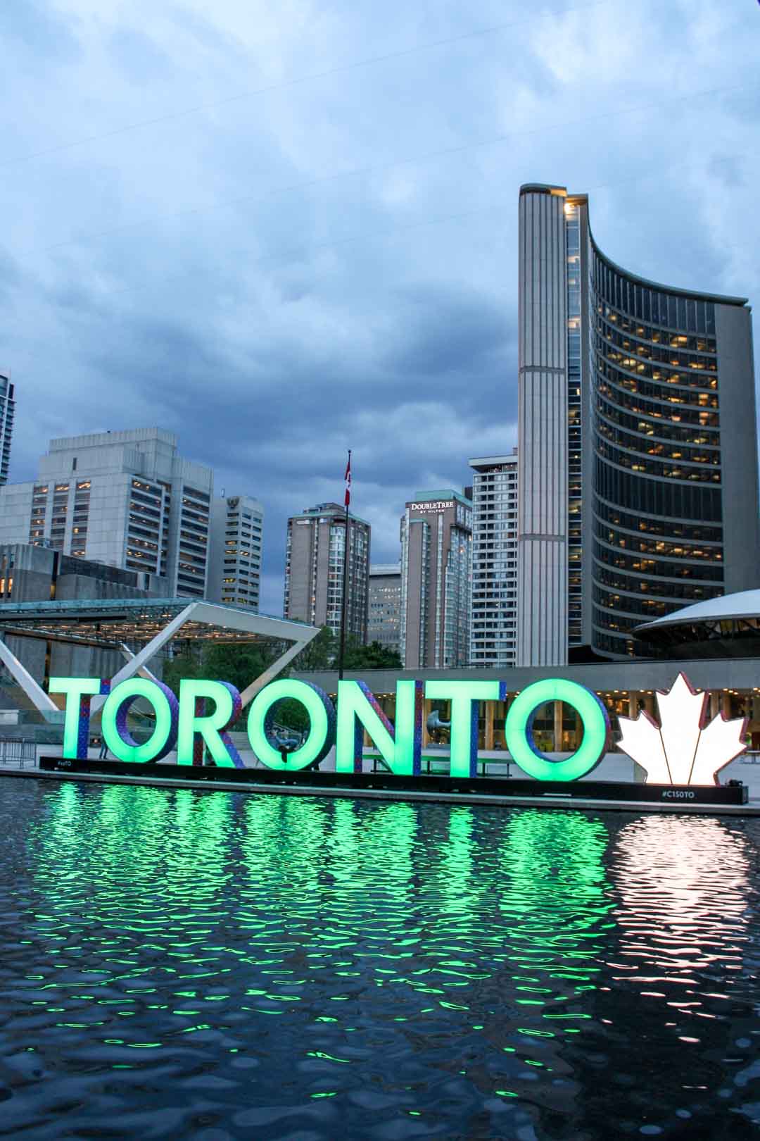 3-D Toronto Sign illuminated in green with Maple Leaf sign illuminated in white as dark clouds fill sky above Toronto skyline