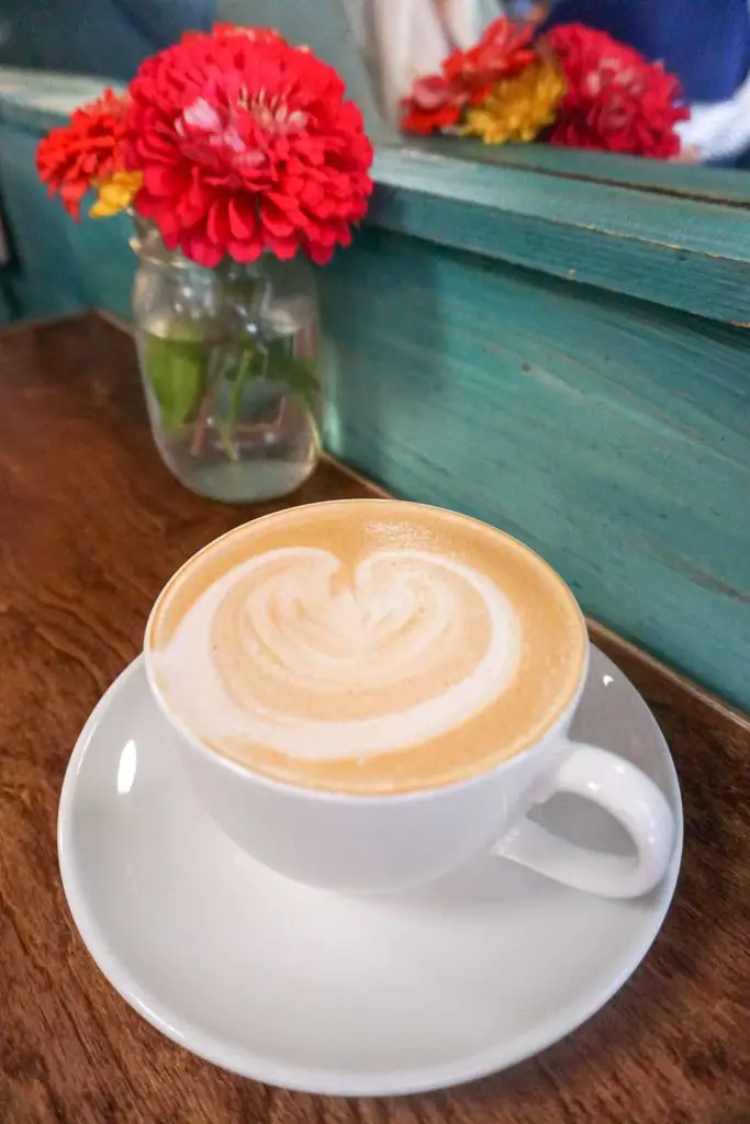 Latte in a white cup and saucer on a wooden table with a mason jar filled with red and yellow flowers at Juicy Kitchen in Ann Arbor, Michigan