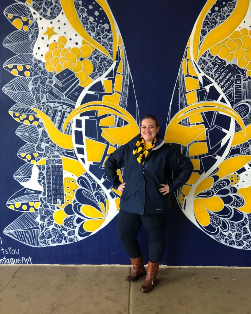 A Michigan fan dressed in maize-and-blue game-day apparel poses in front of the Michigan Wings mural by Kelsey Montague in Ann Arbor, Michigan, USA