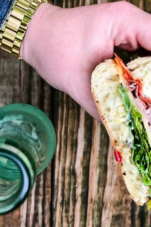 Muffuletta sandwich held in woman's hand by glass bottle with wooden background