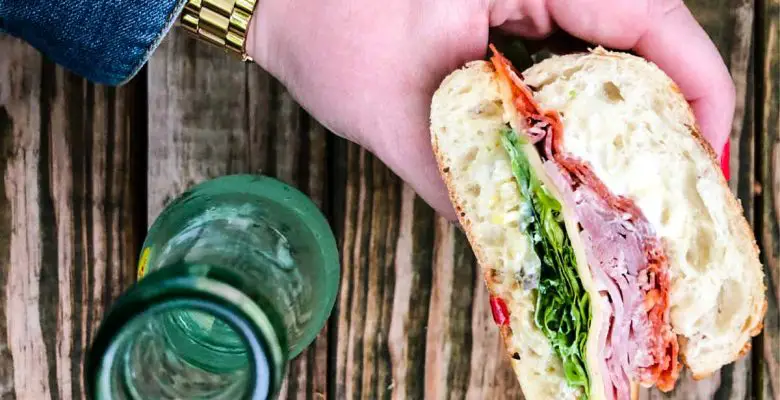 Muffuletta sandwich held in woman's hand by glass bottle with wooden background