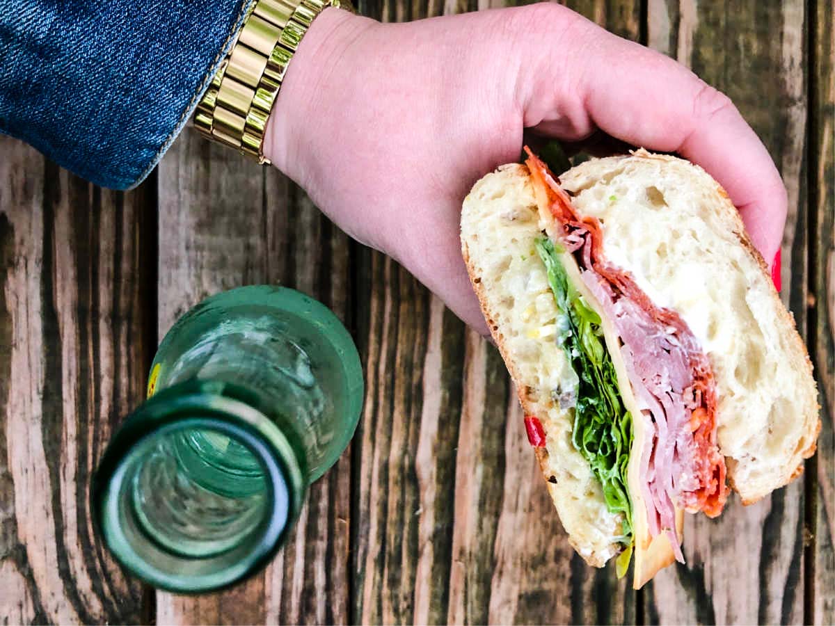 Muffuletta sandwich held in woman's hand by glass bottle with wooden background