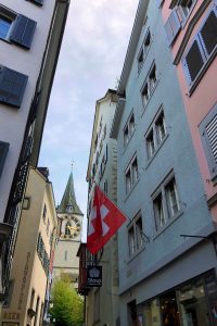 Peter's Church clock peeking through alleyway of colorful buildings displaying Swiss flag