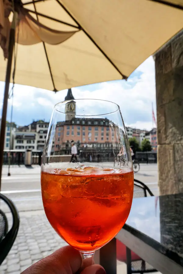 Hand holding Aperol Spritz under tan umbrella with view of Zurich, Switzerland