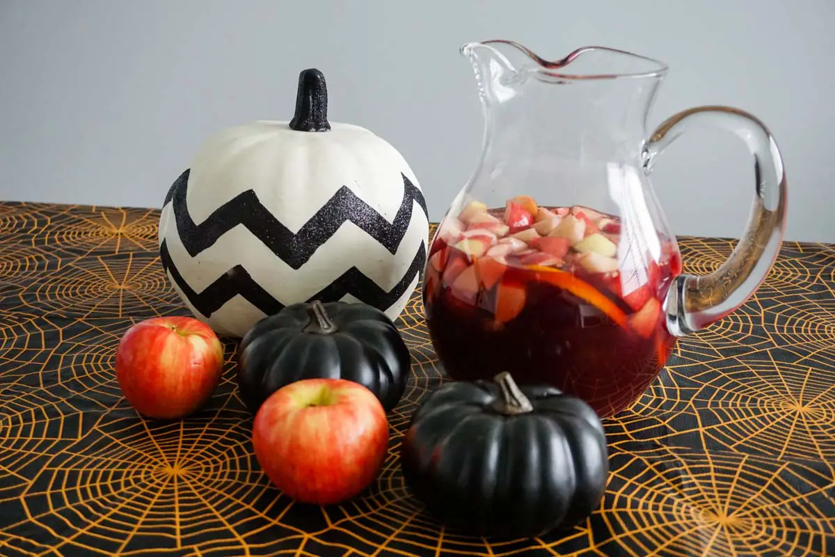 Michigan Apple Cider Sangria with honeycrisp apples in glass potbelly pitcher with assorted decor pumpkins and honeycrisp apples atop Halloween-themed black-and-orange spiderweb tablecloth