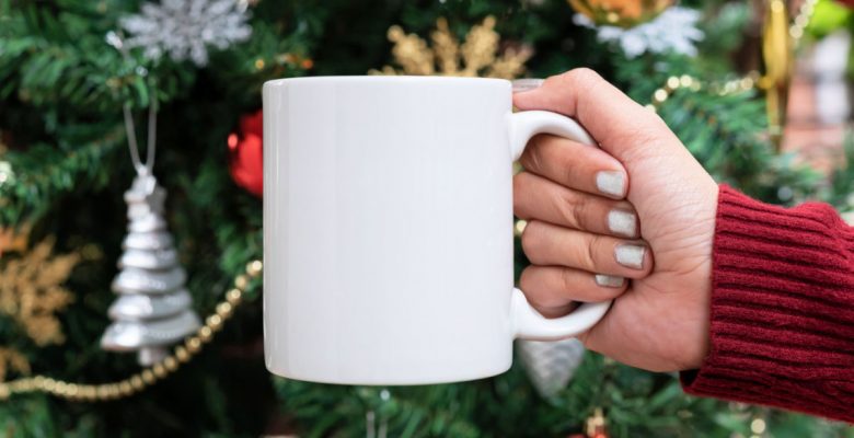 Woman holds white ceramic coffee mug in front of Christmas tree