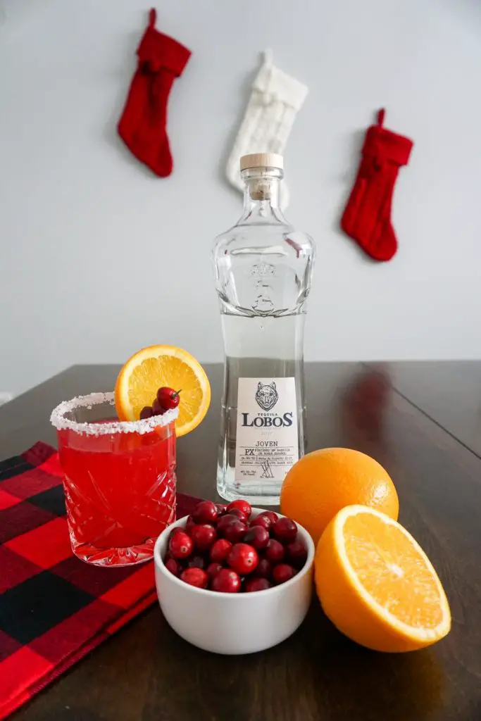 Lobos 1707 Tequila, Joven bottle sits on a dark wooden table with small white bowl of fresh cranberries, halved navel orange, and a cranberry orange margarita in a double old fashioned cocktail glass with a salted rim and fresh cranberry and orange slice garnish in festive holiday setting with red buffalo check cloth and red and white cable knit stockings hanging in background