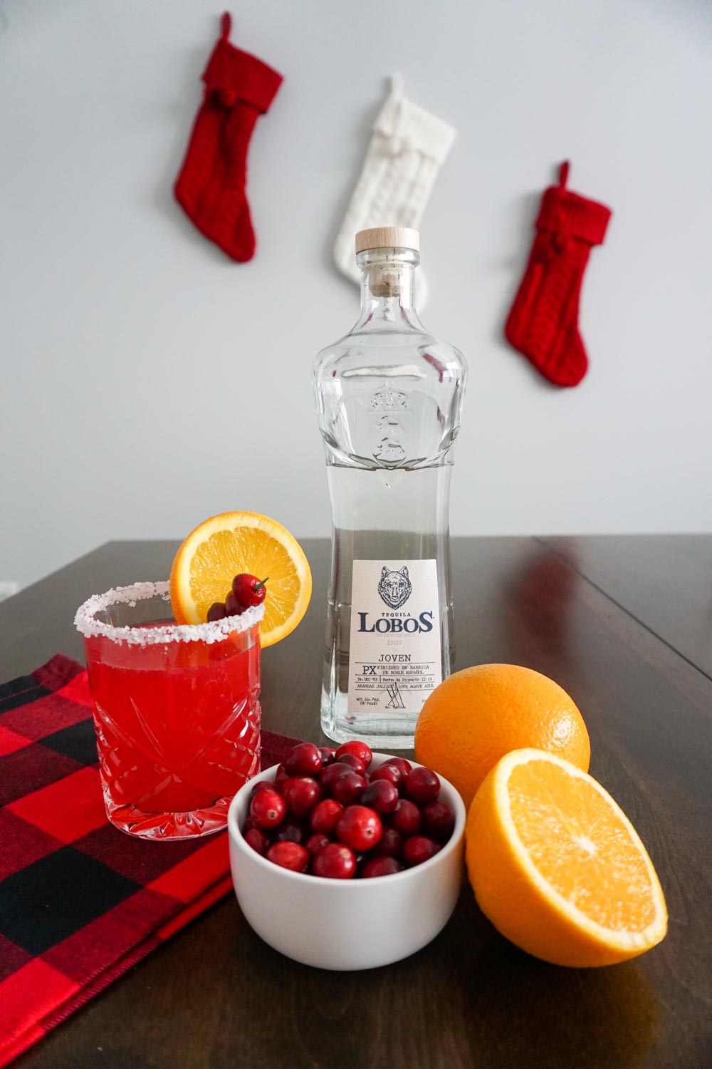 Lobos 1707 Tequila, Joven bottle sits on a dark wooden table with small white bowl of fresh cranberries, halved navel orange, and a cranberry orange margarita in a double old fashioned cocktail glass with a salted rim and fresh cranberry and orange slice garnish in festive holiday setting with red buffalo check cloth and red and white cable knit stockings hanging in background
