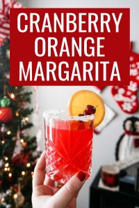 Cranberry Orange Margarita recipe pin showcasing holiday cocktail held in woman's hand in front of a Christmas tree and holiday decorations