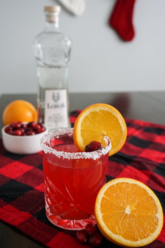 Holiday cranberry orange margarita in festive setting with navel oranges, fresh cranberries, red buffalo check tablecloth, Lobos 1707 Tequila bottle, and Christmas stockings hanging in the background