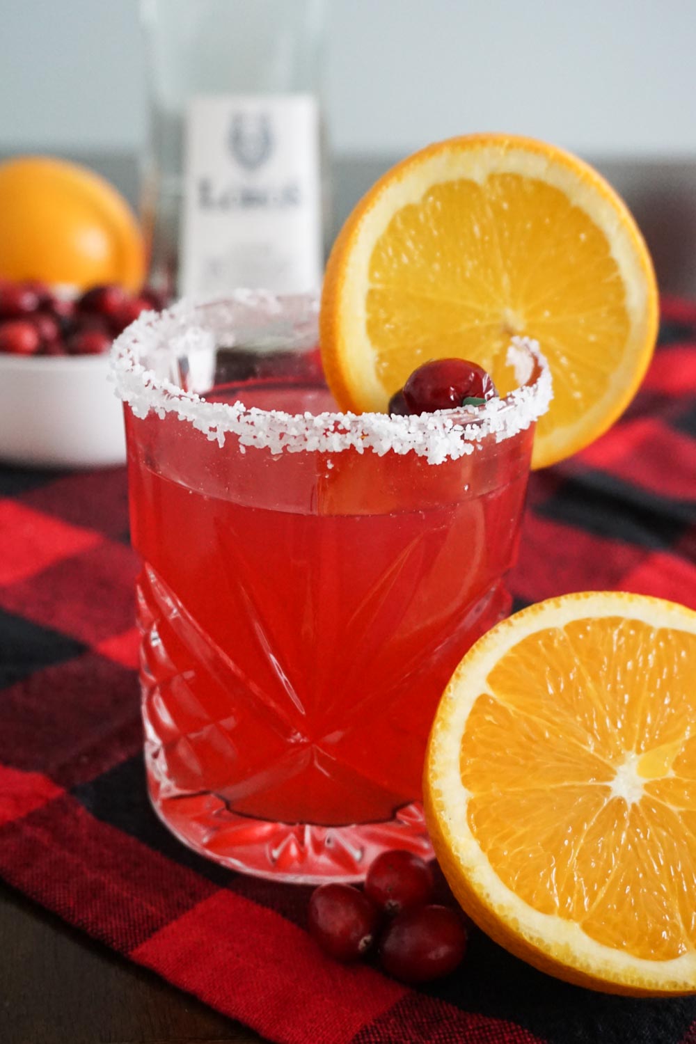 Cranberry orange margarita made with Lobos 1707 Tequila, Joven (pictured in background) in a festive Christmasy setting with red buffalo check tablecloth, navel oranges, and fresh cranberries