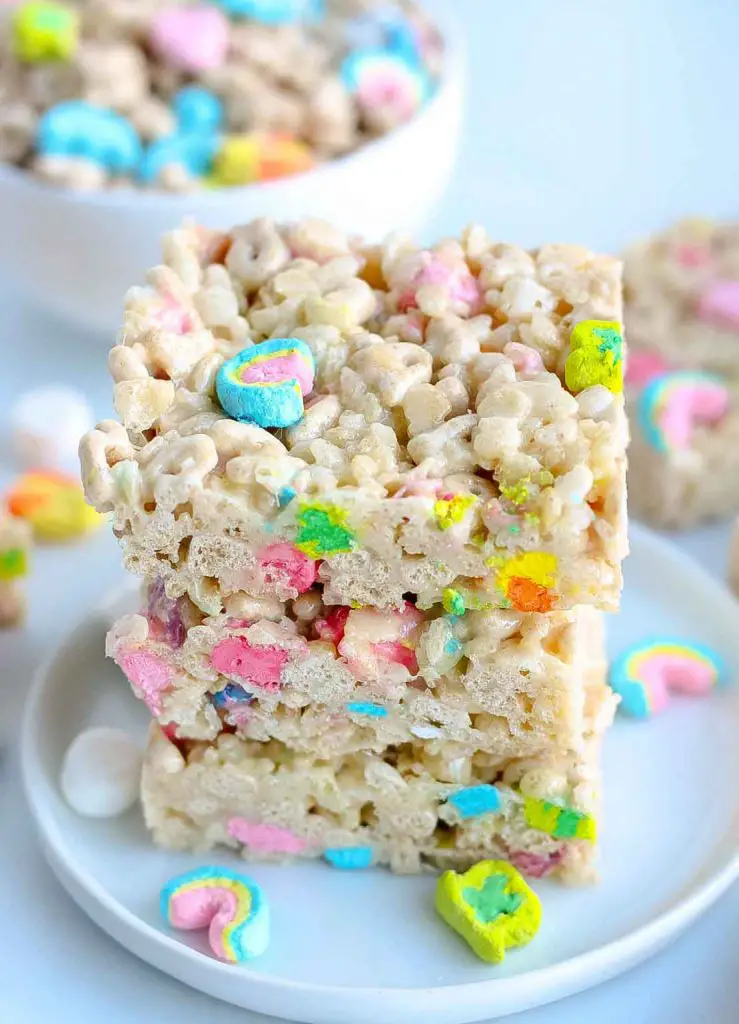Lucky Charms Rice Krispies treats on white plate with bowl of Lucky Charms cereal in background