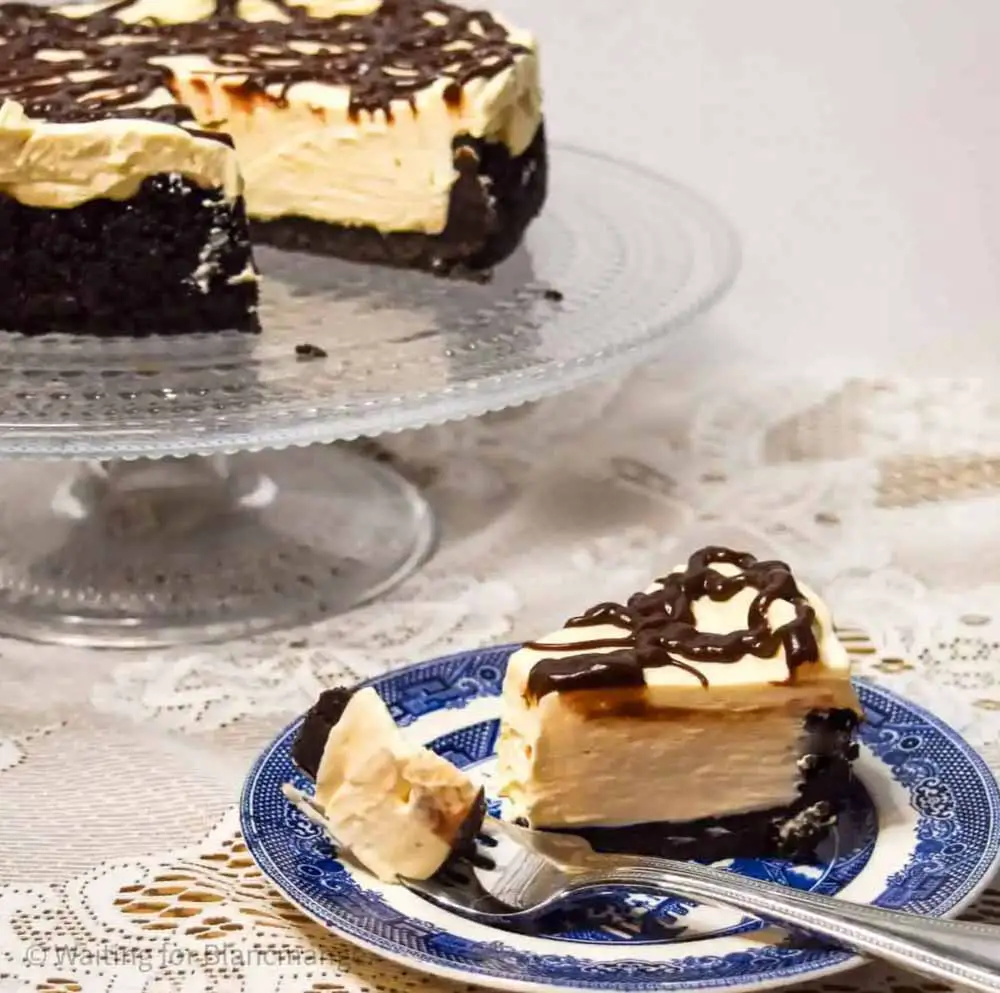 Slice of no-bake Baileys cheesecake on blue plate with remaining cheesecake atop glass cake platter in background