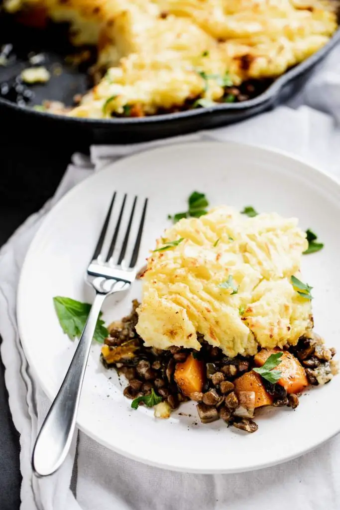 Portion of vegetarian lentil shepherd's pie on a white plate with a fork and pan with remaining shepherd's pie in the background