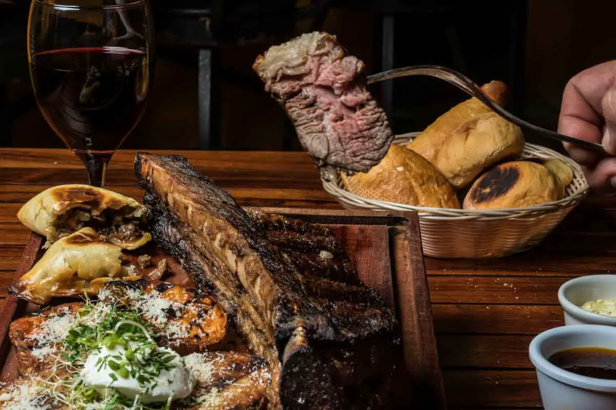 Argenitian meal of asado (grilled meat) plated with an empanada, vegetables, bread basket, dipping sauces, and red wine on a wooden table in a dark setting