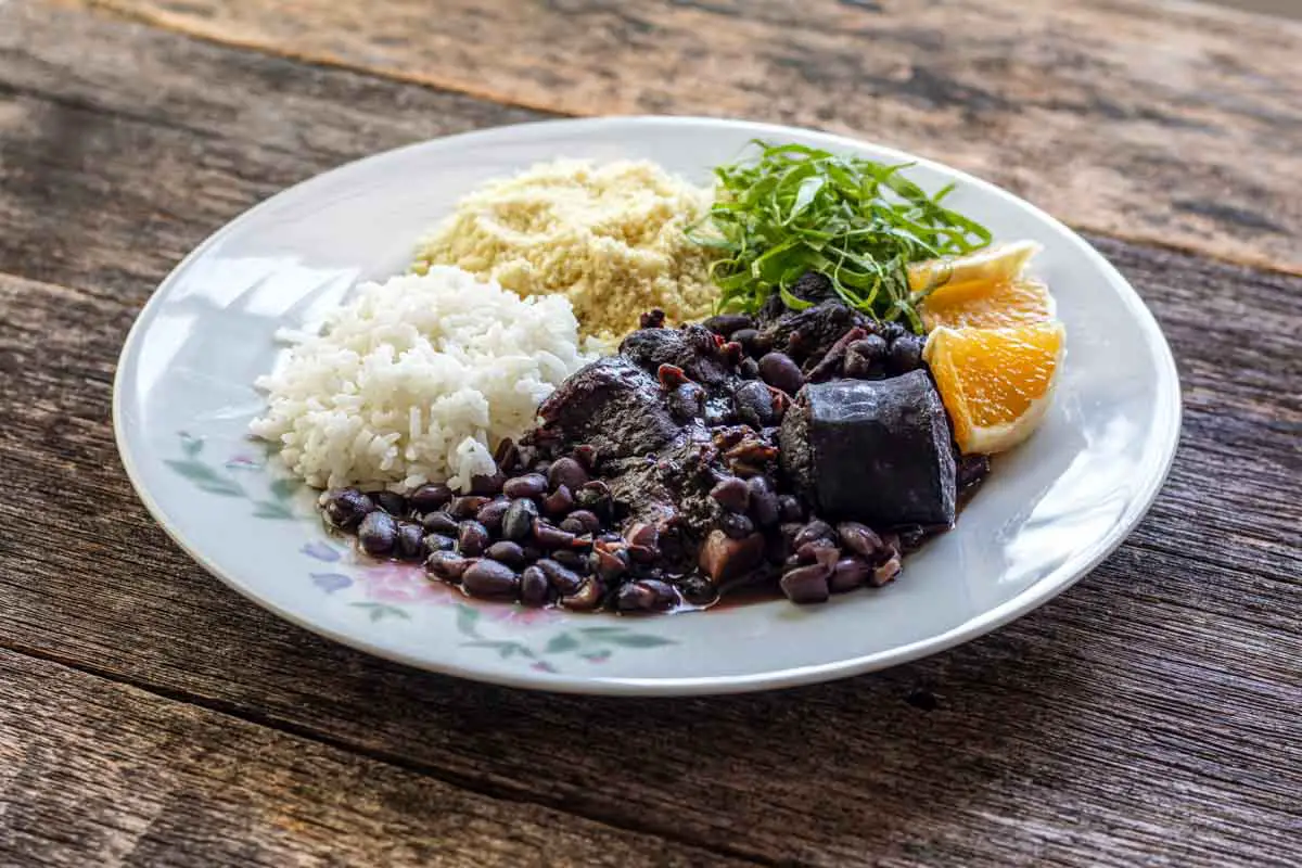 A round white plate with a floral print holds the Brazilian dish feijoada, a black bean stew with pork and beef, alongside rice, orange slices, and greens on a wooden background