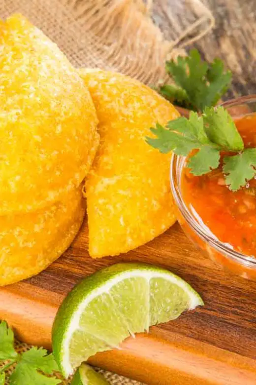 Wooden platter holds six Colombian empanadas (similar to meat pies) with a small glass bowl of red salsa garnished with cilantro leaves and lime wedges on a wooden background