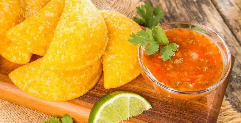 Wooden platter holds six Colombian empanadas (similar to meat pies) with a small glass bowl of red salsa garnished with cilantro leaves and lime wedges on a wooden background