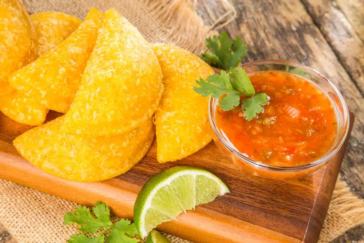 Wooden platter holds six Colombian empanadas (similar to meat pies) with a small glass bowl of red salsa garnished with cilantro leaves and lime wedges on a wooden background
