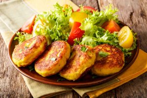 Ecuadoran potato pancakes called llapingachos sit alongside a salad of green lettuce and red and yellow tomatoes on a brown plate sitting atop a folded cloth on a wooden table
