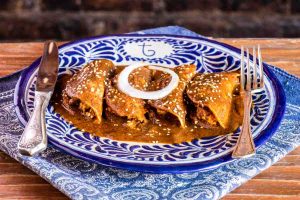 Four Mexican mole enchiladas on a blue-and-white, patterned plate with fork and knife on a wooden table with a dark brick background