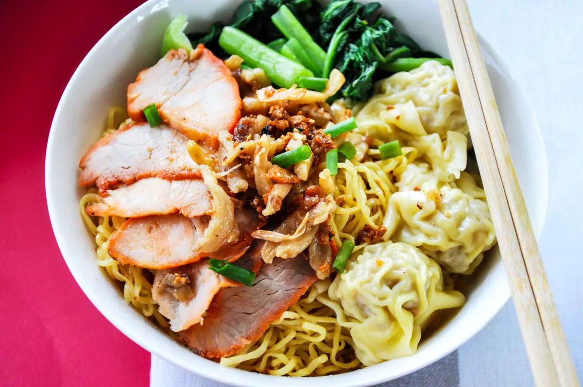 Wanton mee dish of egg noodles, sliced pork, and wantons in a white bowl with chopsticks