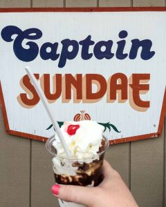 "Famous" Tommy Turtle hot fudge sundae in clear plastic cup held by woman's hand in front of Captain Sundae sign in Holland, Michigan