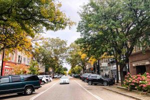 A white car drives down 8th Street lined with shops, parked cars, and trees with leaves changing colors from green to yellow in early fall in downtown Holland, Michigan, USA