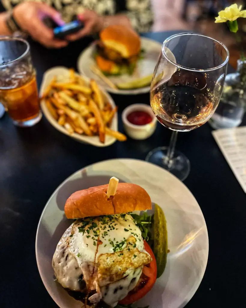 Dark dining table set for two diners with chorizo cheeseburger and glass of rosé wine in foreground and a burger, side of fries with ketchup, and iced tea sit in the background