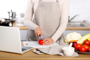 Woman cuts a tomato while watching a cooking tutorial on a laptop