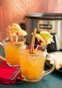 Wassail mulled wine cocktails with sliced apple and cinnamon sticks garnishes with slow cooker in the background