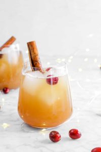 Apple cider bourbon fizz cocktail in stemless glasses with cinnamon stick and fresh cranberries as garnishes in white setting with white string lights