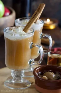 Hot apple pie cocktails topped with whipped cream and cinnamon stick garnish in cozy indoor setting