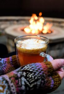 Cup of hot buttered apple cider held in front of a fire pit