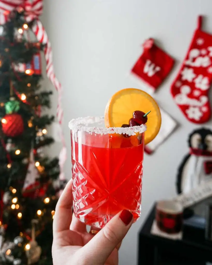 Cranberry orange margarita held in a woman's hand in front of a festive Christmas tree and holiday decorations