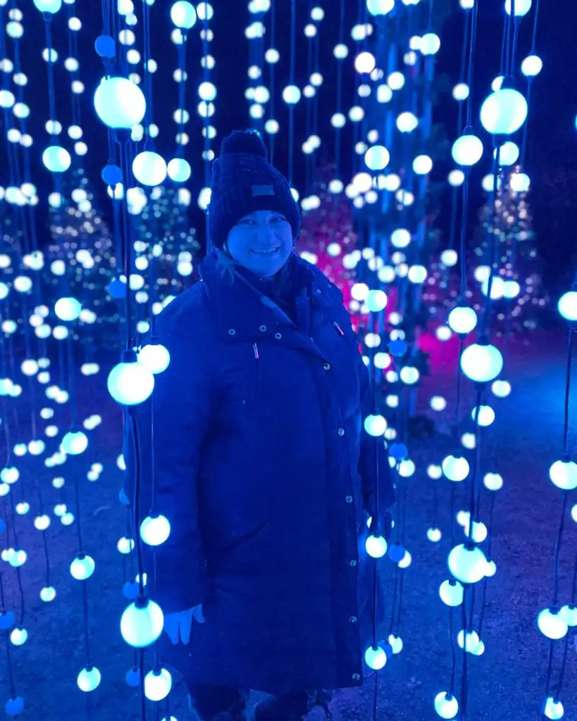 Woman in winter coat and hat poses in blue bubble lights at a holiday illuminated forest trail walk during the holiday season