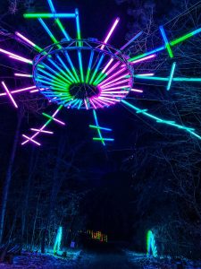 A neon snowflake light display hangs above the trail in the illuminated forest of Glenlore Trails in Commerce Township, Michigan, USA
